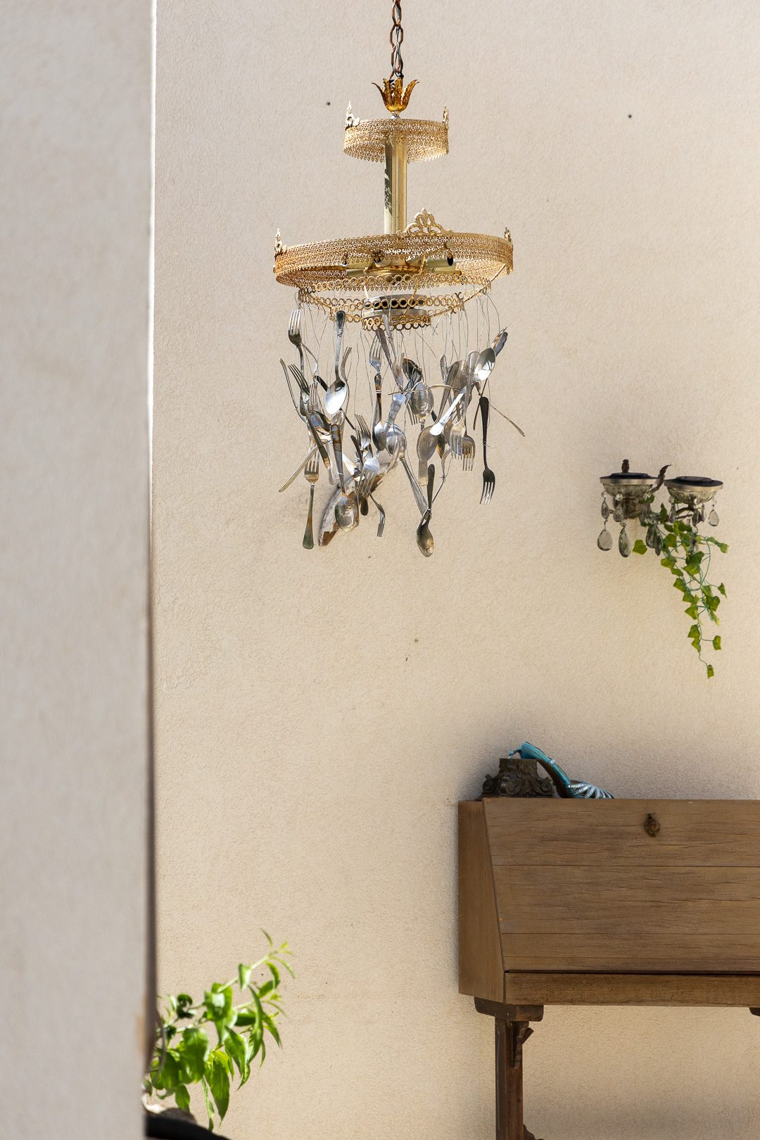 A chandelier made from suspended silver forks and spoons hanging on a sunlit cream wall