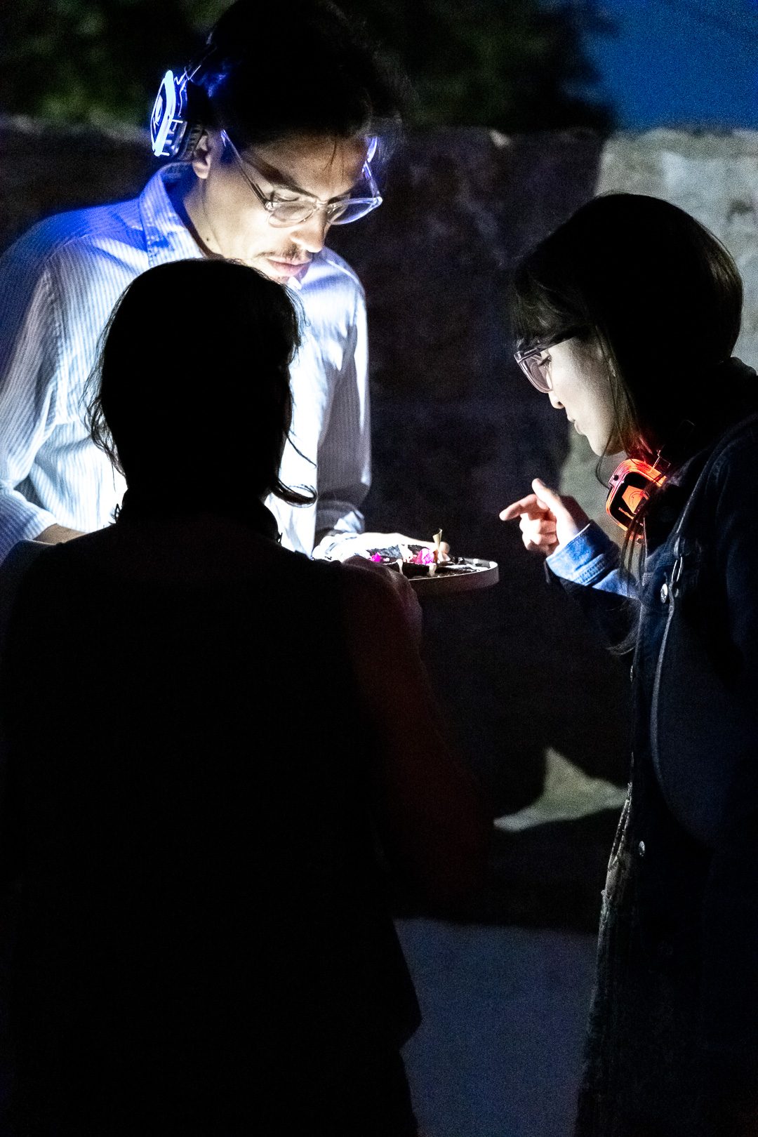 Guests wearing glowing wireless headphones leaning in over a course being served after dark