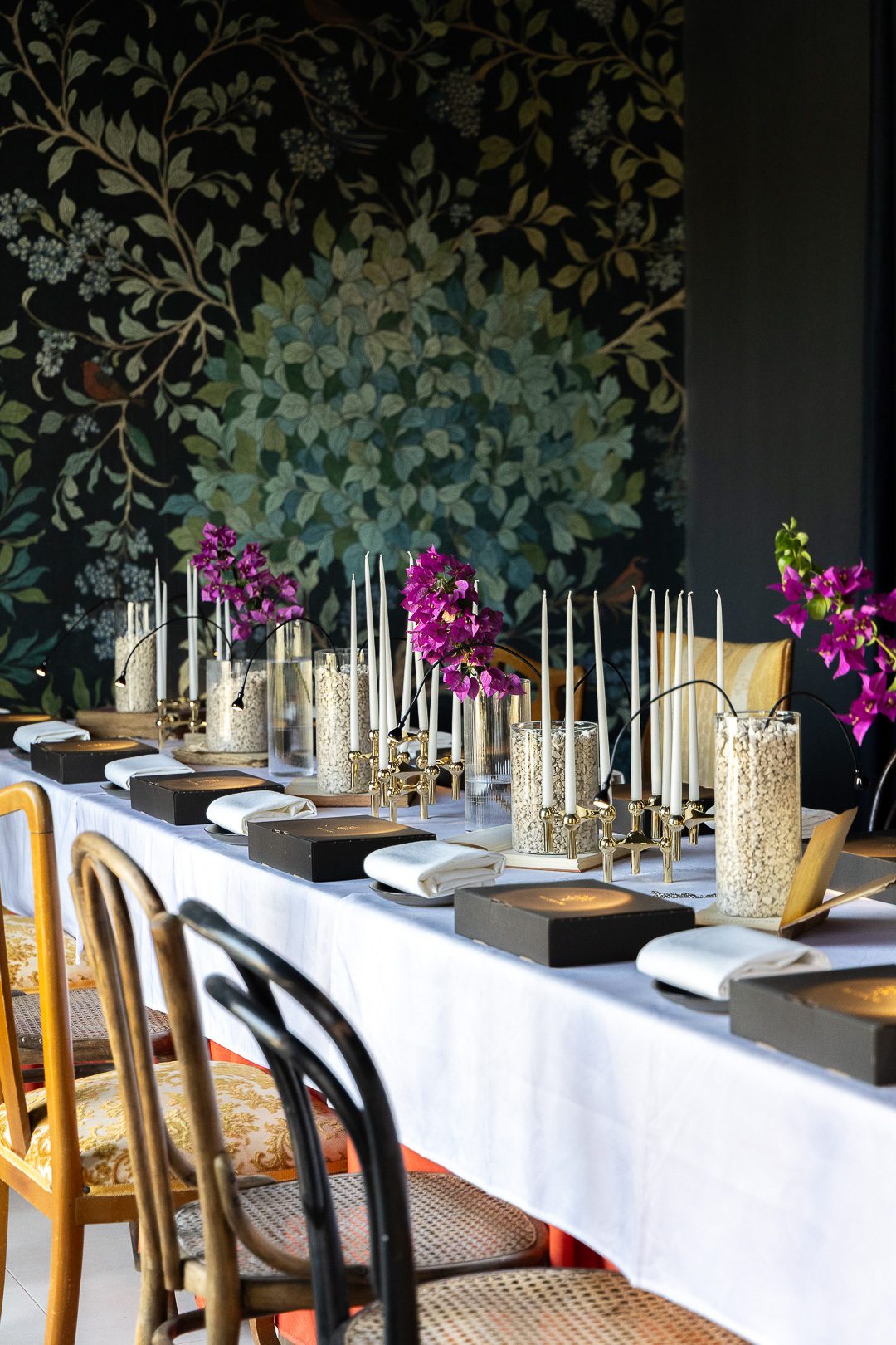 A long table laid in daylight with white linen, tapers and bougainvillea against a dark botanical mural