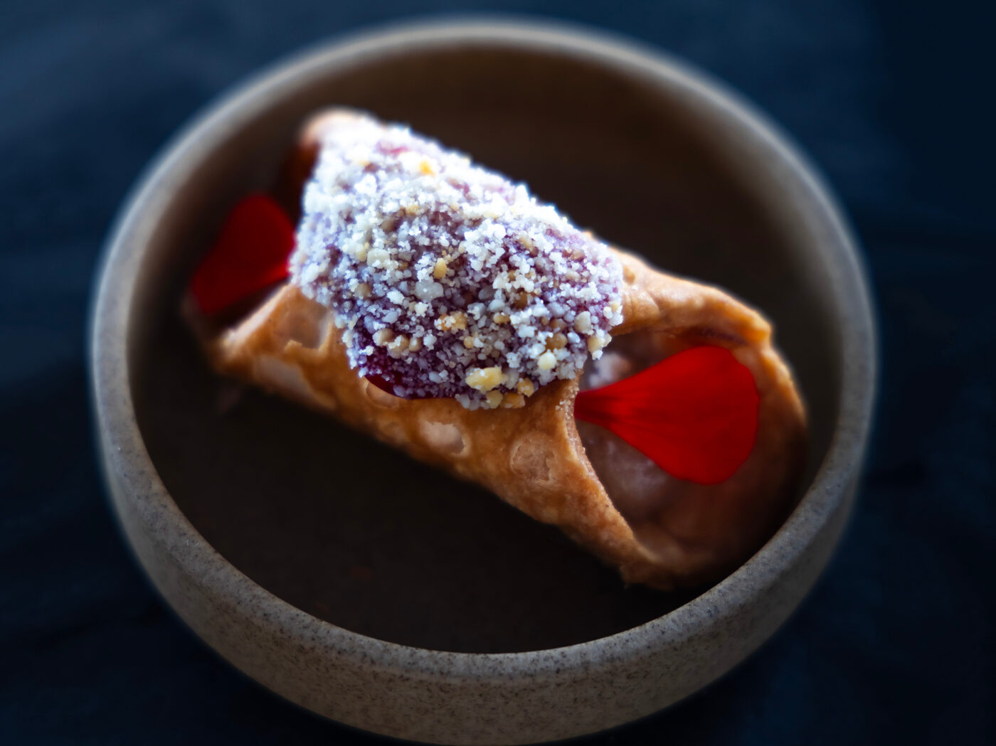 A small cannolo dusted with almond and sugar, scattered with red petals in a stone bowl
