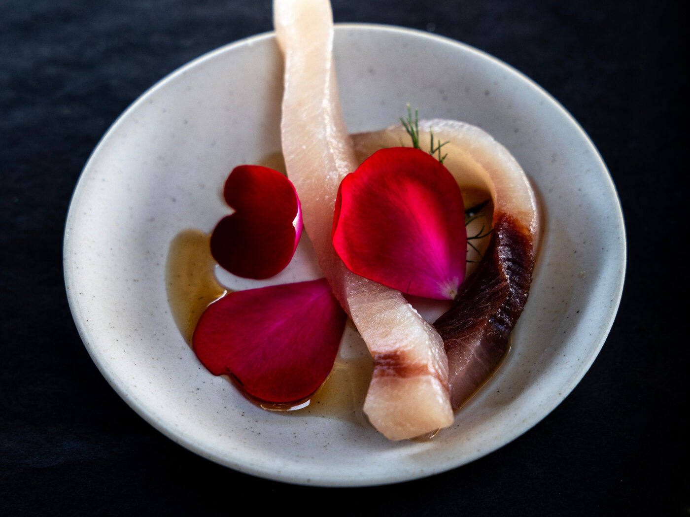 Slices of cured fish with rose petals, fennel fronds and olive oil in a pale stoneware bowl