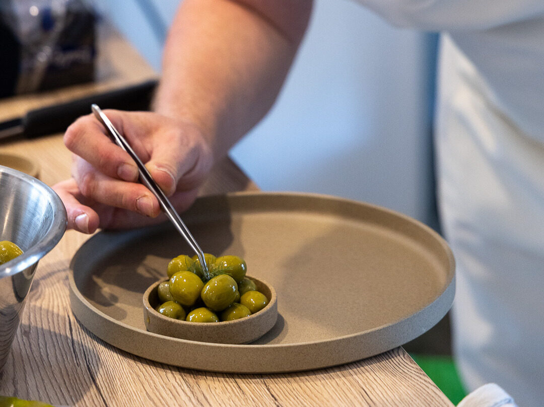 A cook placing green olives one by one into a small stoneware dish with tweezers