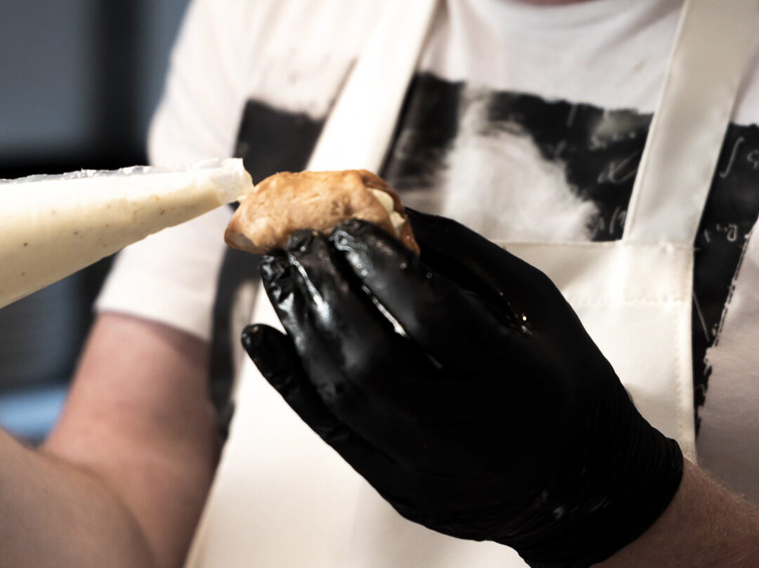 A gloved hand piping cream into a small choux bun in the Five Seasons kitchen