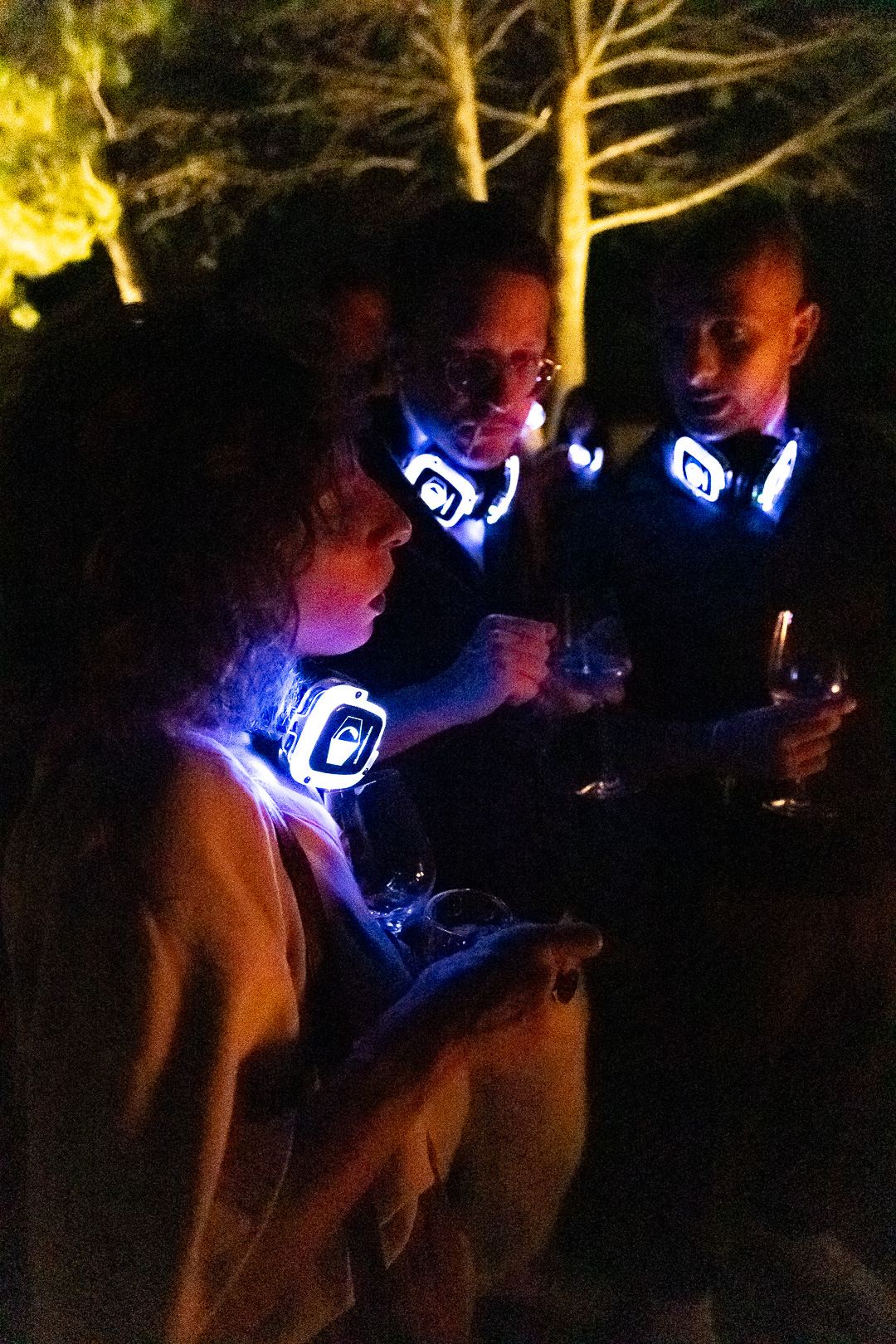 Guests standing in the garden after dark with glasses in hand and glowing headphones around their necks