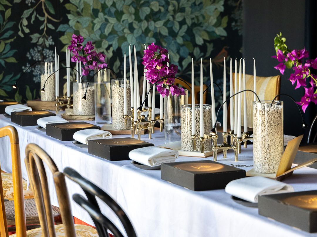 A long table laid in daylight with white linen, tapers and bougainvillea against a green botanical mural