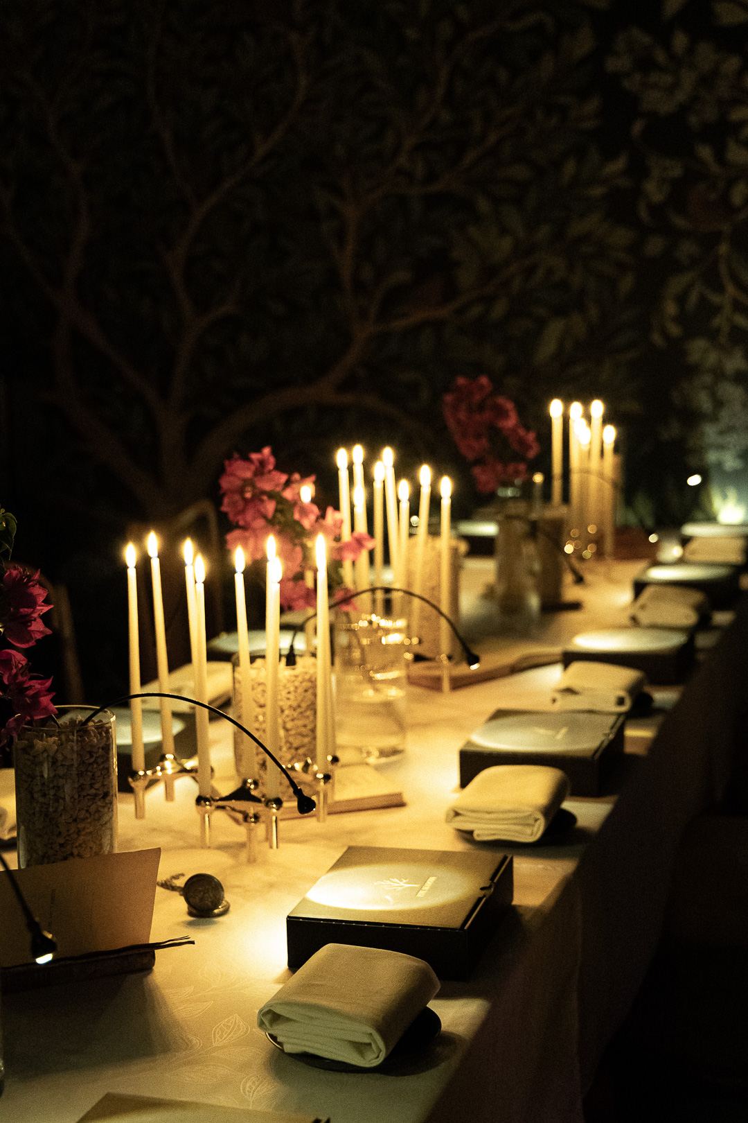 Tapers burning the length of a laid table against a dark botanical mural, with linen and sealed boxes at each place