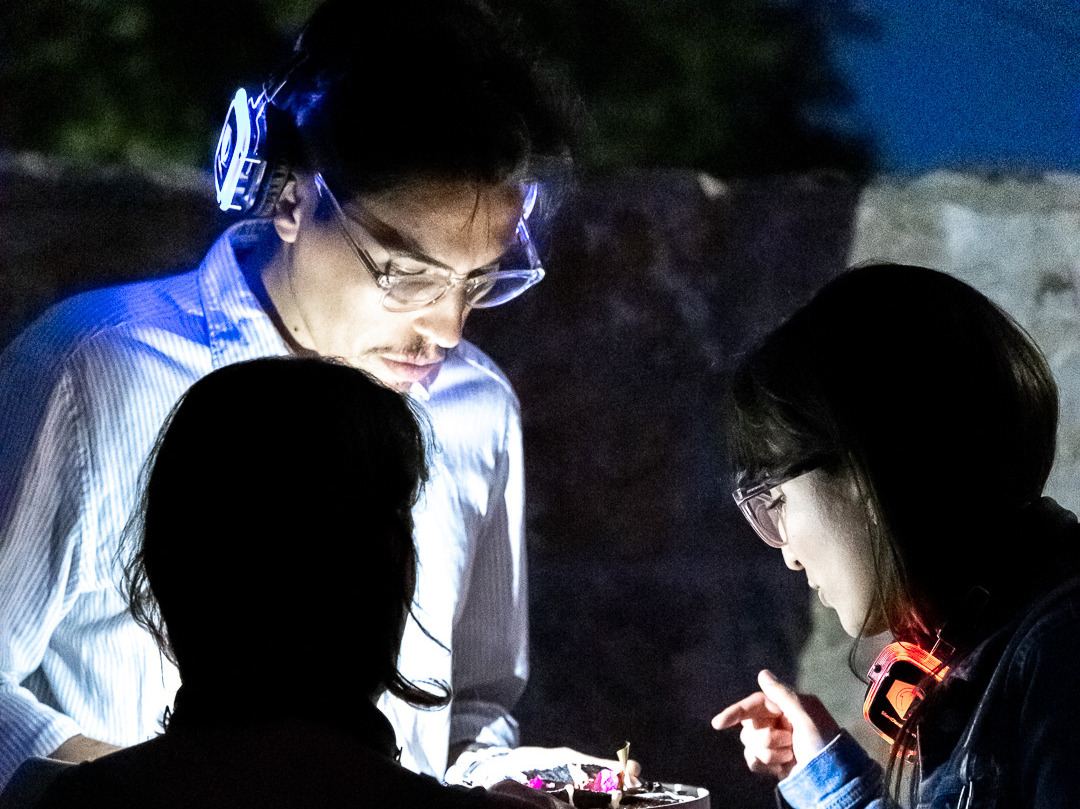 Guests wearing softly glowing wireless headphones leaning in over a course served in the dark