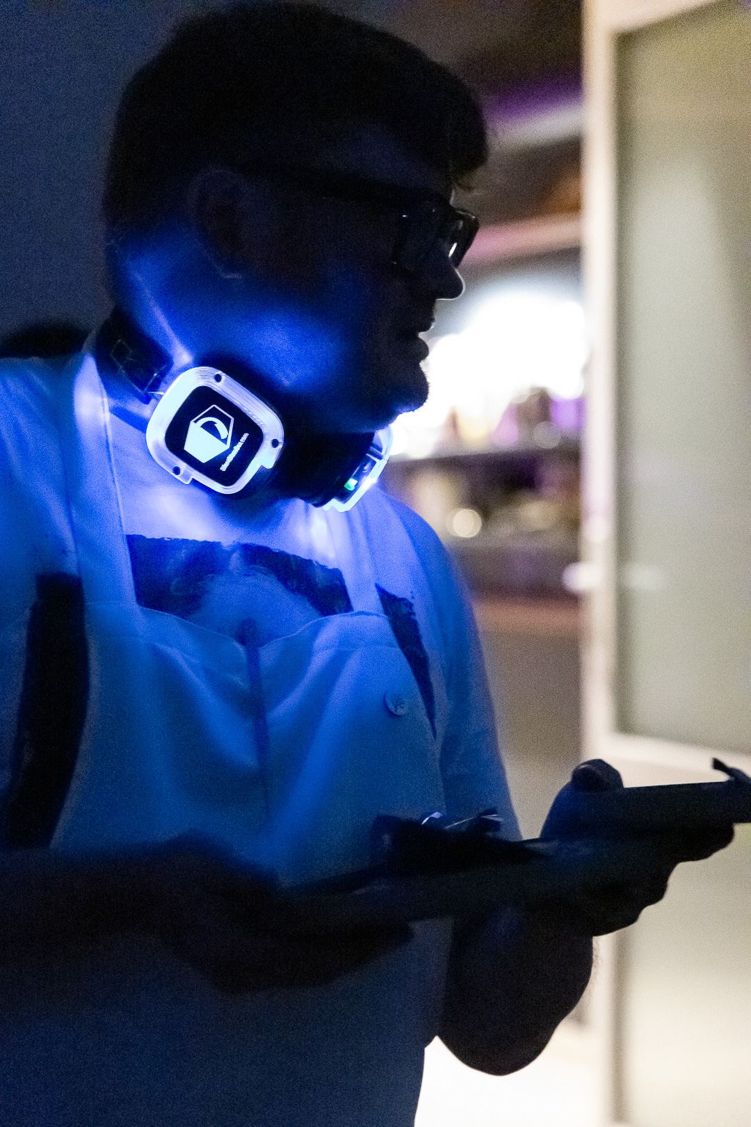 A cook in an apron and headphones carrying a plate through blue light during service